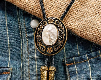 Könnte beinhalten: Ein verzierter Bolo-Krawatte mit einem zentralen ovalen Stein mit rissiger Optik, eingebettet in ein schwarzes und goldenes Filigran-Design. Die Krawatte hat eine schwarze Lederschnur und dekorative goldfarbene Spitzen, vor einer Jeansjacke.