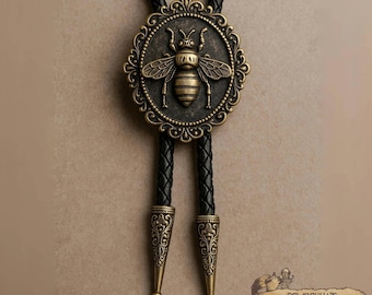 Puede incluir: Una corbata bolo de color bronce con un motivo de abeja detallado dentro de un marco circular ornamentado. La corbata tiene un cordón de cuero trenzado negro con puntas metálicas decorativas. El diseño es de inspiración vintage, mide unos 40 cm.