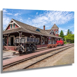 Puede incluir: Una antigua estación de tren con exterior rojo y techo marrón. Un carro de madera está sobre las vías frente a la estación. Un vagón de tren rojo está sobre las vías detrás del carro. El cielo es azul con nubes blancas.
