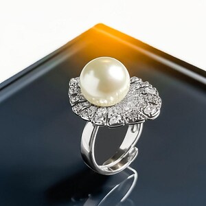 May include: A silver-toned ring featuring a large, round, cream-colored pearl centered atop a fan-shaped setting. The fan-shaped base is encrusted with numerous small, clear, sparkling stones that surround the base of the pearl, creating a bright, reflective, and detailed piece of jewelry.