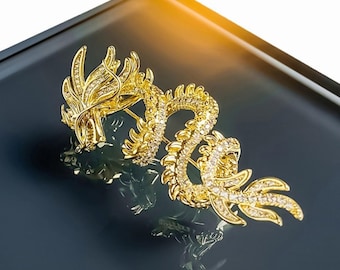 Puede incluir: Broche dorado con forma de dragón, adornado con brillantes cristales. El dragón presenta detalles intrincados, con un cuerpo curvado y una cola fluida. Se muestra sobre una superficie oscura y reflectante.