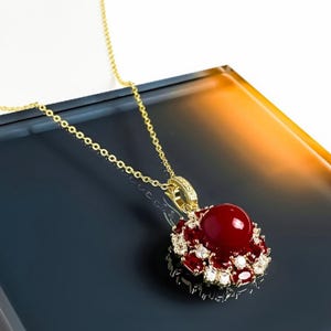 May include: A gold-colored necklace with a pendant featuring a large, round, deep red gemstone at the center, surrounded by smaller red and clear stones. The necklace chain is delicate and the pendant is set on a dark surface.