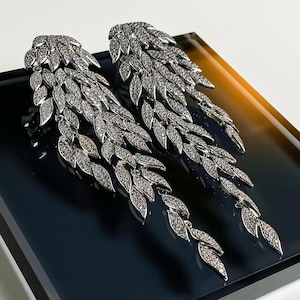 May include: A pair of long, silver-toned dangle earrings featuring a cascading leaf design. Each earring is composed of multiple articulated metallic leaf-shaped segments, entirely encrusted with small, clear, sparkling stones that catch the light.