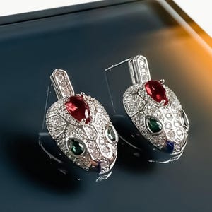 May include: A pair of silver-toned snake-shaped earrings. Each earring features a red teardrop-shaped stone at the top, with green and white stones forming the eyes and head. The snake's head is encrusted with small, clear stones.