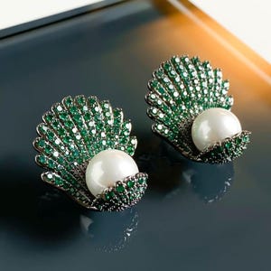 May include: A pair of earrings with a shell design, each holding a white pearl. The shells are adorned with green gems and set in a silver-tone metal. The earrings are displayed against a dark backdrop, accentuating the details.