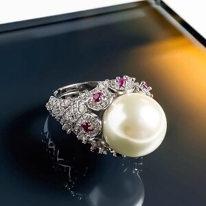 May include: A silver-toned ring featuring a large, round, white pearl as the centerpiece. The band is intricately decorated with small, clear, sparkling stones and several small, round, vibrant red rubies set in a textured, ornate metal design.