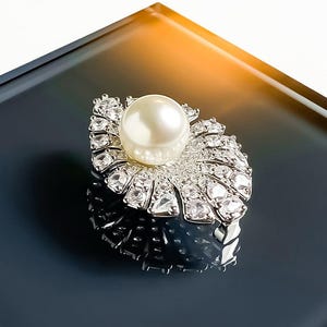 May include: A silver-toned brooch featuring a central round white pearl surrounded by a fan-shaped arrangement of clear, faceted, sparkling stones. The piece has a metallic base with a polished finish and a reflective surface texture.