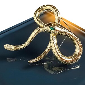 May include: A gold-toned snake brooch featuring a textured scale pattern, a single marquise-cut green stone for the eye, and small clear crystals embedded along the tail section. The coiled serpent design is polished to a high shine finish.