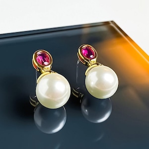 Könnte beinhalten: Ein Paar goldfarbene Ohrringe mit einer runden weißen Perle, die an einem kleinen, ovalen, leuchtend pinken Edelstein in einer polierten goldfarbenen Metallfassung hängt. Die Ohrringe haben eine glatte, reflektierende Oberfläche.