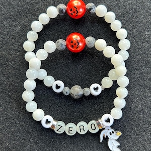 May include: Three beaded bracelets. The top two bracelets have white beads, gray beads, and red beads with a jack-o'-lantern face. The bottom bracelet has white letter beads spelling "ZERO" and a charm of a ghost dog.