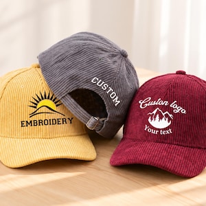 Può includere: Tre cappellini da baseball in velluto a coste nei colori giallo, grigio e bordeaux. Il cappellino giallo presenta una grafica con sole e montagna e la scritta EMBROIDERY. Il cappellino grigio riporta la parola CUSTOM. Quello bordeaux ha un logo.