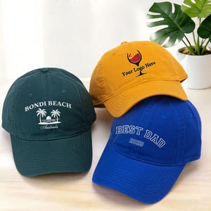 Puede incluir: Tres gorras de béisbol apiladas. Una es verde azulado con "Bondi Beach Australia" bordado. Otra es amarillo mostaza con un logotipo de copa de vino y "Your Logo Here". La tercera es azul con "BEST DAD 2025".