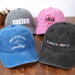 May include: Four baseball caps in grey, pink, blue, and black. The grey cap features the word custom. The pink cap has a red heart and the word mom. The blue cap displays Paul & Shark yachting with a shark graphic. The black cap says Embroidery.