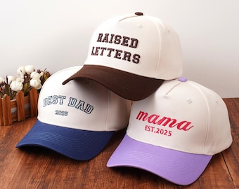 May include: Three baseball caps with white crowns and colored brims. One cap has a brown brim and the words "RAISED LETTERS" in brown. Another has a blue brim and the words "BEST DAD 2025" in blue. The third has a purple brim and the word "mama EST.2025" in pink.