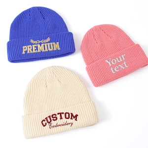 May include: Three ribbed knit beanies in blue, pink, and cream. The blue beanie has "PREMIUM" embroidered in gold. The pink beanie has "Your text" embroidered in silver. The cream beanie has "CUSTOM Embroidery" embroidered in burgundy.