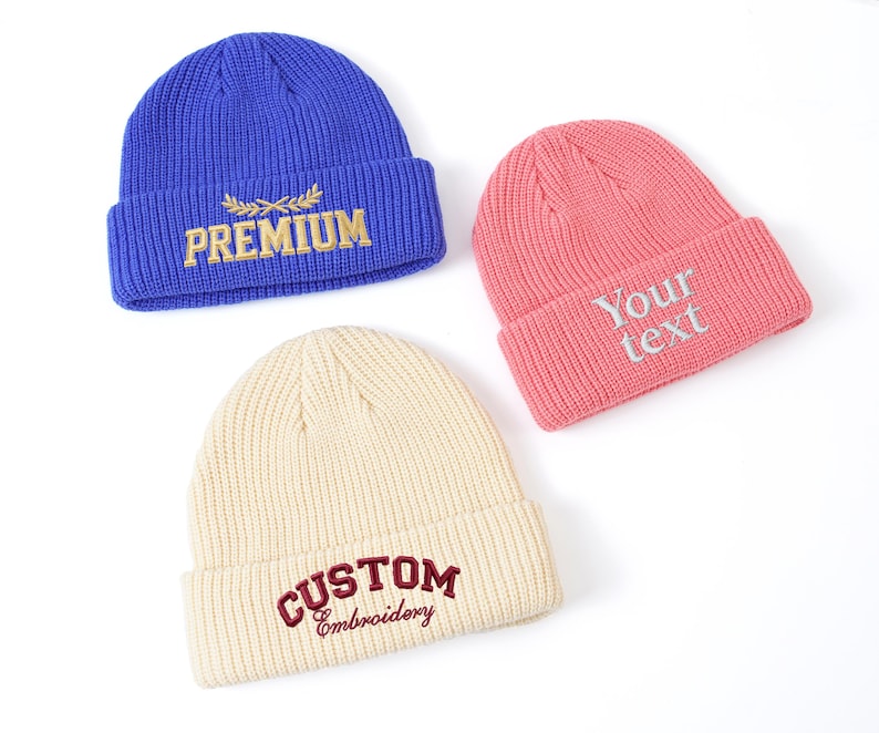 May include: Three ribbed knit beanies in blue, pink, and cream. The blue beanie has "PREMIUM" embroidered in gold. The pink beanie has "Your text" embroidered in silver. The cream beanie has "CUSTOM Embroidery" embroidered in burgundy.