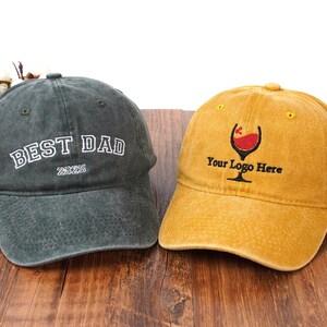 Pu&ograve; includere: Un cappellino da baseball grigio antracite con testo ricamato in bianco BEST DAD 2025 e un cappellino da baseball giallo senape con un grafico ricamato di un calice da vino e il testo nero Your Logo Here, entrambi con finitura in tessuto usurato.