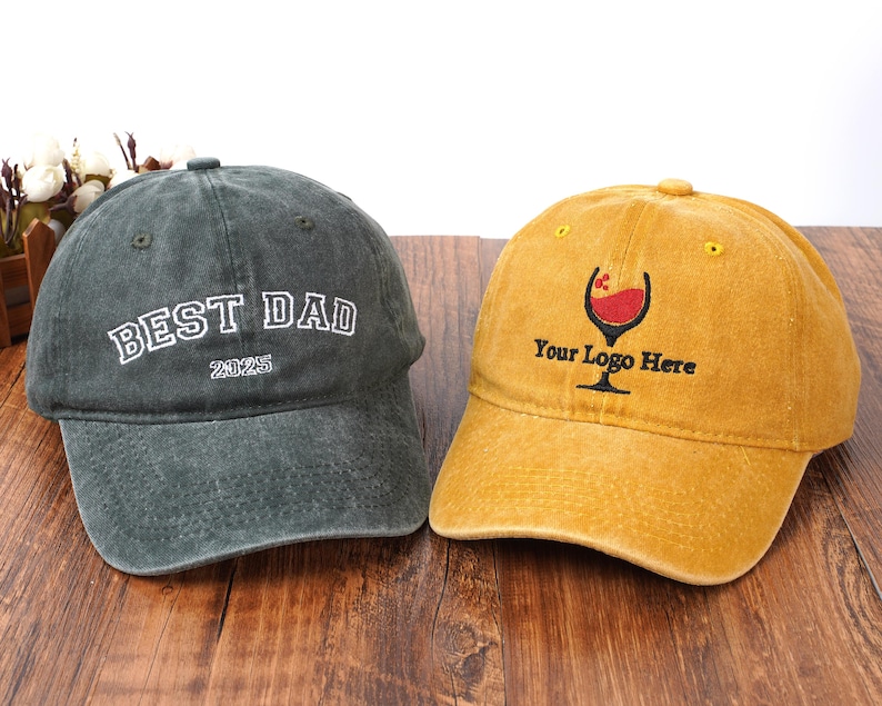 Pu&ograve; includere: Un cappellino da baseball grigio antracite con testo ricamato in bianco BEST DAD 2025 e un cappellino da baseball giallo senape con un grafico ricamato di un calice da vino e il testo nero Your Logo Here, entrambi con finitura in tessuto usurato.