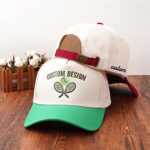 Puede incluir: Dos gorras de béisbol blancas con viseras verdes. Una gorra tiene bordado "CUSTOM DESIGN" con raquetas de tenis y una pelota. La otra gorra tiene bordado "custom". Ambas tienen correas burdeos.