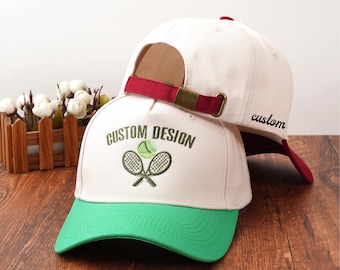 May include: Two white baseball caps with green visors. One cap has "CUSTOM DESIGN" embroidered with tennis rackets and a ball. The other cap has "custom" embroidered on the side. Both have burgundy straps.