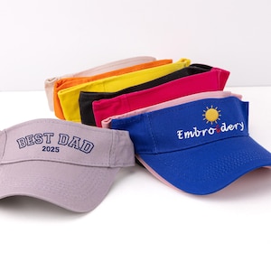 K&ouml;nnte beinhalten: Eine Sammlung farbenfroher Sonnenblenden. Ein lavendelfarbenes Visier zeigt "BEST DAD 2025" in Marineblau. Ein blaues Visier hat "Embroidery" und eine Sonnengrafik. Weitere Visiere sind orange, gelb, schwarz und rosa.