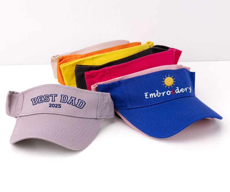 K&ouml;nnte beinhalten: Eine Sammlung farbenfroher Sonnenblenden. Ein lavendelfarbenes Visier zeigt "BEST DAD 2025" in Marineblau. Ein blaues Visier hat "Embroidery" und eine Sonnengrafik. Weitere Visiere sind orange, gelb, schwarz und rosa.