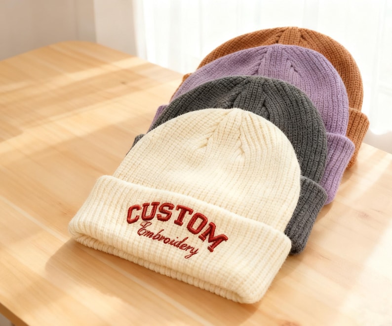 May include: A stack of ribbed knit beanies in various colors: cream, gray, purple, and brown. The cream beanie features the embroidered text "CUSTOM Embroidery" in red. The hats are arranged on a light wood surface.