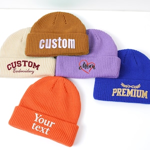 May include: A collection of six beanies in various colors, including orange, brown, purple, cream, and blue. Each beanie features embroidered text such as "custom", "mom", "premium", and "Your text". The beanies are displayed on a white surface.