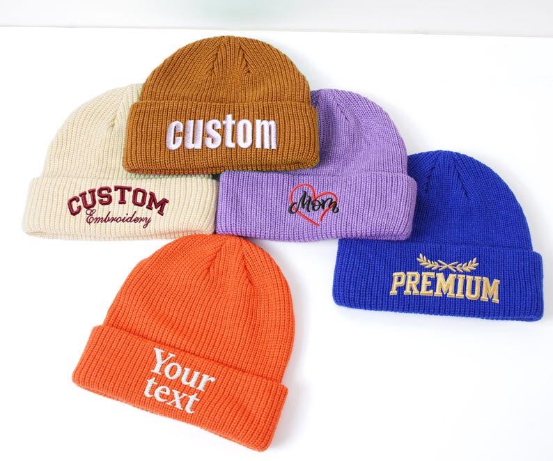 May include: A collection of six beanies in various colors, including orange, brown, purple, cream, and blue. Each beanie features embroidered text such as "custom", "mom", "premium", and "Your text". The beanies are displayed on a white surface.