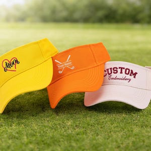 K&ouml;nnte beinhalten: Drei Golfvisiere in Gelb, Orange und Wei&szlig; werden auf gr&uuml;nem Gras gezeigt. Das gelbe Visier hat ein Herz und das Wort "Mom". Das orangefarbene Visier hat gekreuzte Golfschl&auml;ger und "Your Name". Das wei&szlig;e Visier sagt "CUSTOM Embroidery".