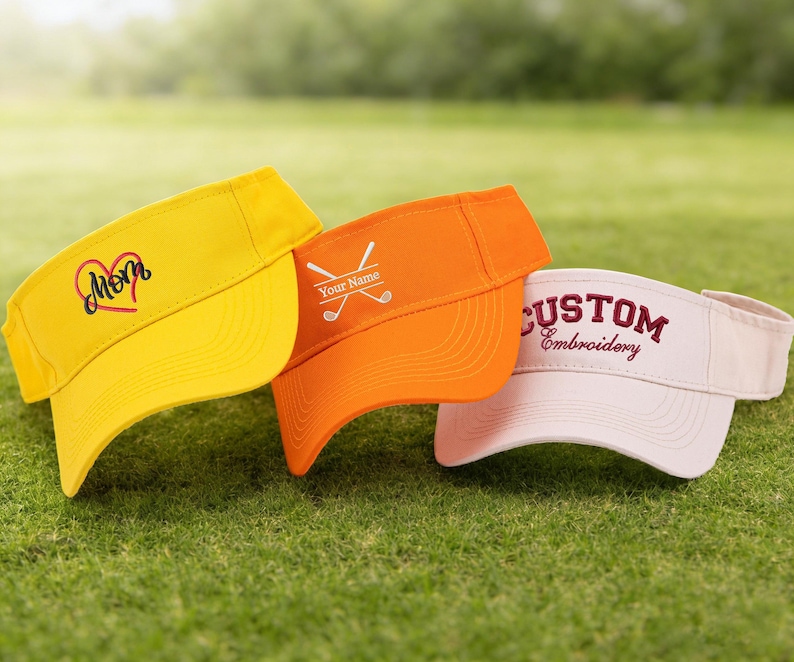 K&ouml;nnte beinhalten: Drei Golfvisiere in Gelb, Orange und Wei&szlig; werden auf gr&uuml;nem Gras gezeigt. Das gelbe Visier hat ein Herz und das Wort "Mom". Das orangefarbene Visier hat gekreuzte Golfschl&auml;ger und "Your Name". Das wei&szlig;e Visier sagt "CUSTOM Embroidery".