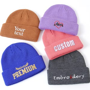 May include: A collection of colorful knit beanies. The beanies are in shades of brown, purple, pink, gray, and blue. Each beanie has embroidered text, including "Your text", "Mom", "Custom", "Premium", and "Embroidery".