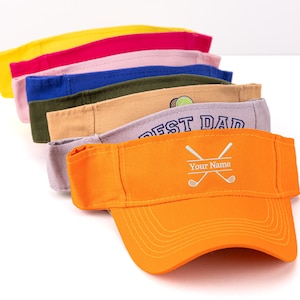 K&ouml;nnte beinhalten: Ein Stapel bunter Sonnenblenden in Gelb, Pink, Blau, Gr&uuml;n, Beige, Grau und Orange. Das orangefarbene Visier hat ein Golfmotiv mit gekreuzten Schl&auml;gern und dem Text "Your Name". Das graue Visier sagt "BEST DAD".