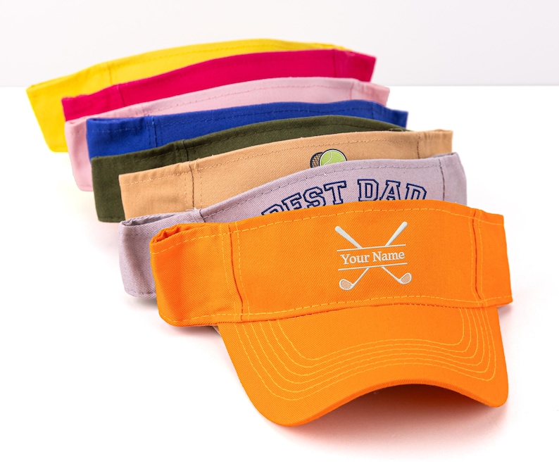 K&ouml;nnte beinhalten: Ein Stapel bunter Sonnenblenden in Gelb, Pink, Blau, Gr&uuml;n, Beige, Grau und Orange. Das orangefarbene Visier hat ein Golfmotiv mit gekreuzten Schl&auml;gern und dem Text "Your Name". Das graue Visier sagt "BEST DAD".