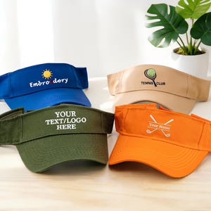 May include: Four visors in blue, tan, green, and orange. The blue visor has a sun and the word "Embroidery". The tan visor says "Tennis Club". The green visor says "YOUR TEXT/LOGO HERE". The orange visor has crossed golf clubs and "Your Name".