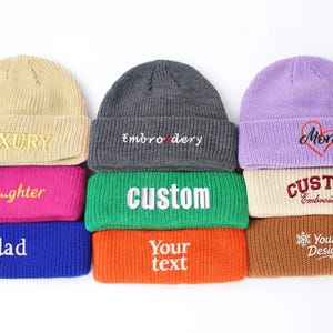 May include: A collection of colorful knit beanies with embroidered text. The beanies are in various colors, including tan, gray, purple, pink, green, blue, and orange. The text includes "LUXURY", "Embroidery", "Mom", "Daughter", "CUSTOM", "dad", and "Your text".