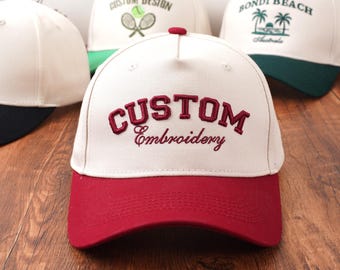 May include: A collection of baseball caps with various designs. The front-most cap is cream-colored with a red brim and the words "CUSTOM Embroidery" embroidered in burgundy. Other caps feature designs like tennis rackets and the words "BONDI BEACH Australia".