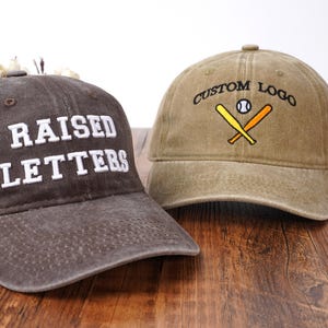 Pu&ograve; includere: Un cappellino da baseball grigio scuro con testo ricamato in bianco RAISED LETTERS e un cappellino da baseball color marrone chiaro con testo ricamato in nero CUSTOM LOGO sopra un'illustrazione di una palla e due mazze incrociate.