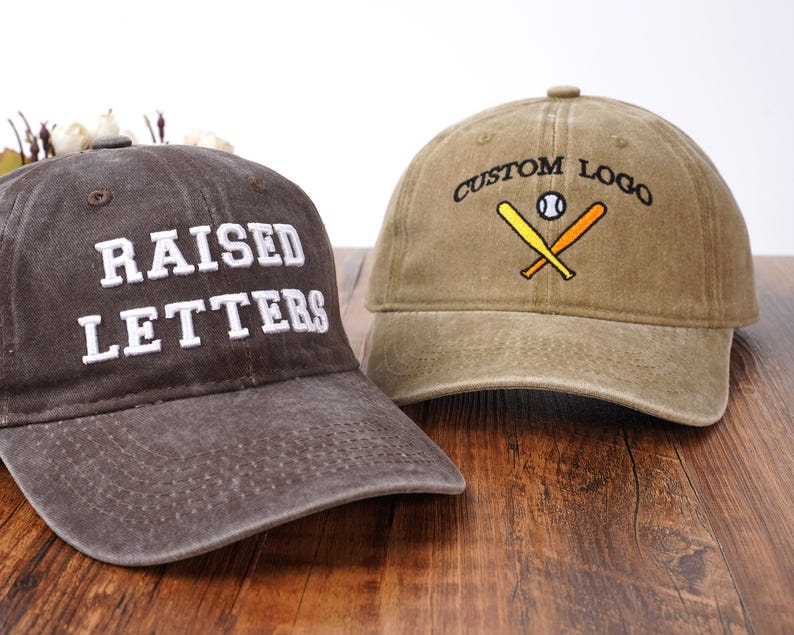 Pu&ograve; includere: Un cappellino da baseball grigio scuro con testo ricamato in bianco RAISED LETTERS e un cappellino da baseball color marrone chiaro con testo ricamato in nero CUSTOM LOGO sopra un'illustrazione di una palla e due mazze incrociate.