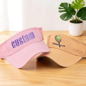 K&ouml;nnte beinhalten: Zwei Sonnenblenden werden gezeigt. Eine ist rosa mit "custom" in Lila gestickt. Die andere ist hellbraun mit einer Tennisball- und Schl&auml;gergrafik und dem Text "Tennis Club". Beide liegen auf einer Holzoberfl&auml;che.