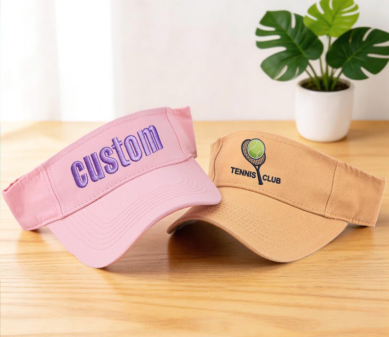 K&ouml;nnte beinhalten: Zwei Sonnenblenden werden gezeigt. Eine ist rosa mit "custom" in Lila gestickt. Die andere ist hellbraun mit einer Tennisball- und Schl&auml;gergrafik und dem Text "Tennis Club". Beide liegen auf einer Holzoberfl&auml;che.