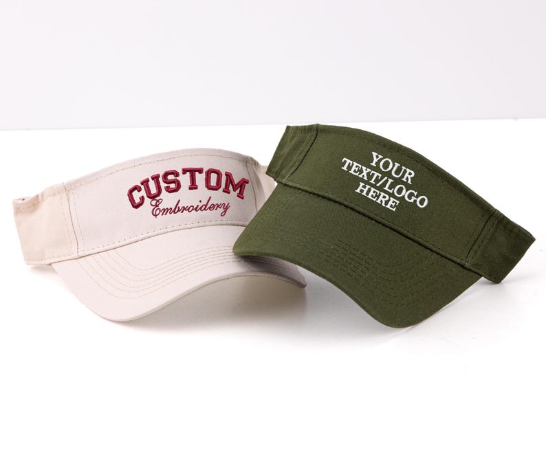 K&ouml;nnte beinhalten: Zwei Sonnenblenden, eine beige und eine olivgr&uuml;ne, werden gezeigt. Die beige Sonnenblende hat die Stickerei "CUSTOM Embroidery" in Burgunderrot. Die gr&uuml;ne Sonnenblende hat "YOUR TEXT/LOGO HERE" in Wei&szlig;. Beide Sonnenblenden sind auf wei&szlig;em Hintergrund.