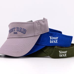 K&ouml;nnte beinhalten: Drei Visore in Grau, Blau und Olivgr&uuml;n sind gestapelt. Auf dem grauen Visor ist "BEST DAD 2025" aufgestickt. Die blauen und gr&uuml;nen Visore tragen den Text "Your text" aufgestickt.