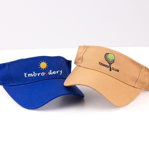 K&ouml;nnte beinhalten: Zwei Sonnenblenden: eine blaue Sonnenblende mit "Embroidery" und einem Sonnenlogo und eine beige Sonnenblende mit einem Tennis-Schl&auml;ger- und Ball-Logo mit der Aufschrift "Tennis Club". Beide Sonnenblenden sind aus robustem Material gefertigt und sollen die Augen vor der Sonne sch&uuml;tzen.