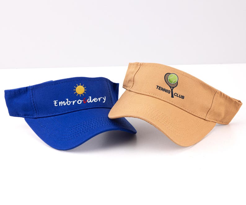 K&ouml;nnte beinhalten: Zwei Sonnenblenden: eine blaue Sonnenblende mit "Embroidery" und einem Sonnenlogo und eine beige Sonnenblende mit einem Tennis-Schl&auml;ger- und Ball-Logo mit der Aufschrift "Tennis Club". Beide Sonnenblenden sind aus robustem Material gefertigt und sollen die Augen vor der Sonne sch&uuml;tzen.