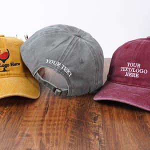 Pu&ograve; includere: Tre cappellini da baseball in giallo senape, grigio e bordeaux. Il cappellino giallo presenta un grafico di un calice di vino e il testo "Your Logo Here". Il grigio ha "YOUR TEXT" ricamato sul lato. Il bordeaux mostra "YOUR TEXT/LOGO HERE".