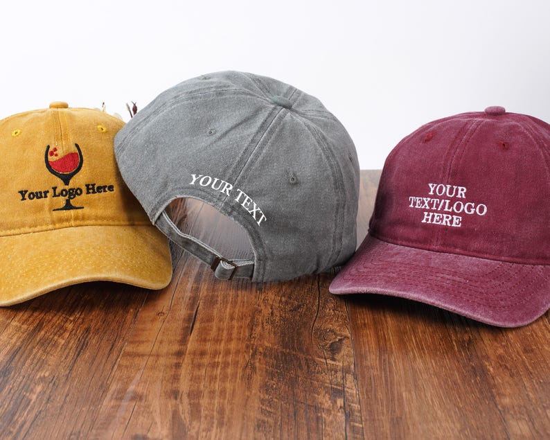 Pu&ograve; includere: Tre cappellini da baseball in giallo senape, grigio e bordeaux. Il cappellino giallo presenta un grafico di un calice di vino e il testo "Your Logo Here". Il grigio ha "YOUR TEXT" ricamato sul lato. Il bordeaux mostra "YOUR TEXT/LOGO HERE".