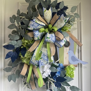 Könnte beinhalten: Ein dekorativer Kranz mit einer zentralen Schleife aus Jute und blauem Hortensien-Musterband, akzentuiert durch lindgrünes Band. Der Kranz enthält künstlichen Eukalyptus, blaue und weiße Hortensienblüten sowie ein hölzernes Vogelhaus mit einem blauen Vogel.