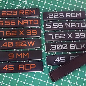 May include: A set of ten black rectangular labels with white and orange text. The labels are arranged in two columns of five. The text on the labels includes various caliber names, such as .223 REM, 5.56 NATO, 7.62 X 39, .40 S&W, .300 BLK, 9 MM, and .45 ACP.