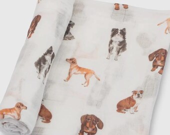 May include: A white muslin blanket featuring various dog breeds in brown, tan, and black. The blanket is rolled up on one side, with the dog illustrations scattered across the fabric. The blanket is made of a soft, lightweight material.